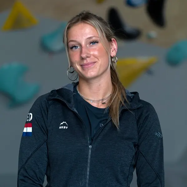 Een vrouw met blond haar in een paardenstaart, met hoepeloorbellen, kettingen en een donker jasje met een patch van de Nederlandse vlag, staat binnenshuis met klimgrepen op de achtergrond, die wazig z